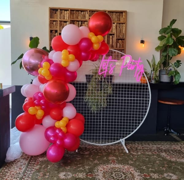 A white mesh frame with a pink and red balloon garland at an indoors party.