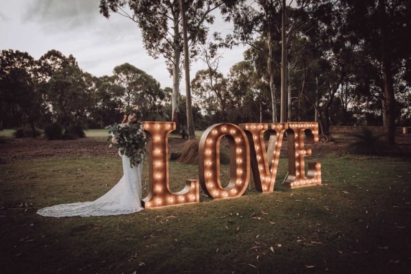 Bride standing next to giant light up love letters at an outdoor wedding.