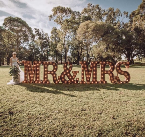Bride posing with giant rustic light up MR & MRS letters in an outdoor setting.