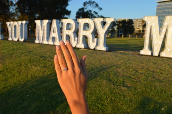Engaged woman holding up her ring in front of giant WILL YOU MARRY ME light up letters.