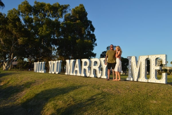Newly engaged couple standing in front of WILL YOU MARRY ME light up letters.