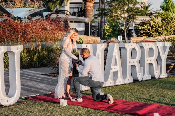 A man proposing on bended knee in front of WILL YOU MARRY ME light up letters.
